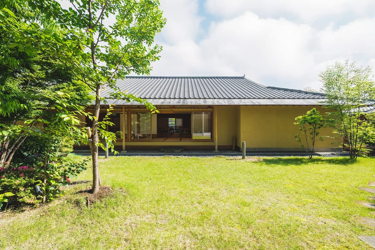 芝生の庭越しに見る瓦屋根の和風建築。木立に囲まれた平屋
